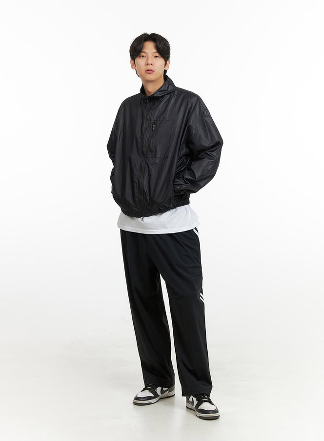 LEWKIN Men's Solid Trackpants IY431 in black, full front view on male model, wide-leg silhouette with white side stripes, paired with black windbreaker and sneakers.