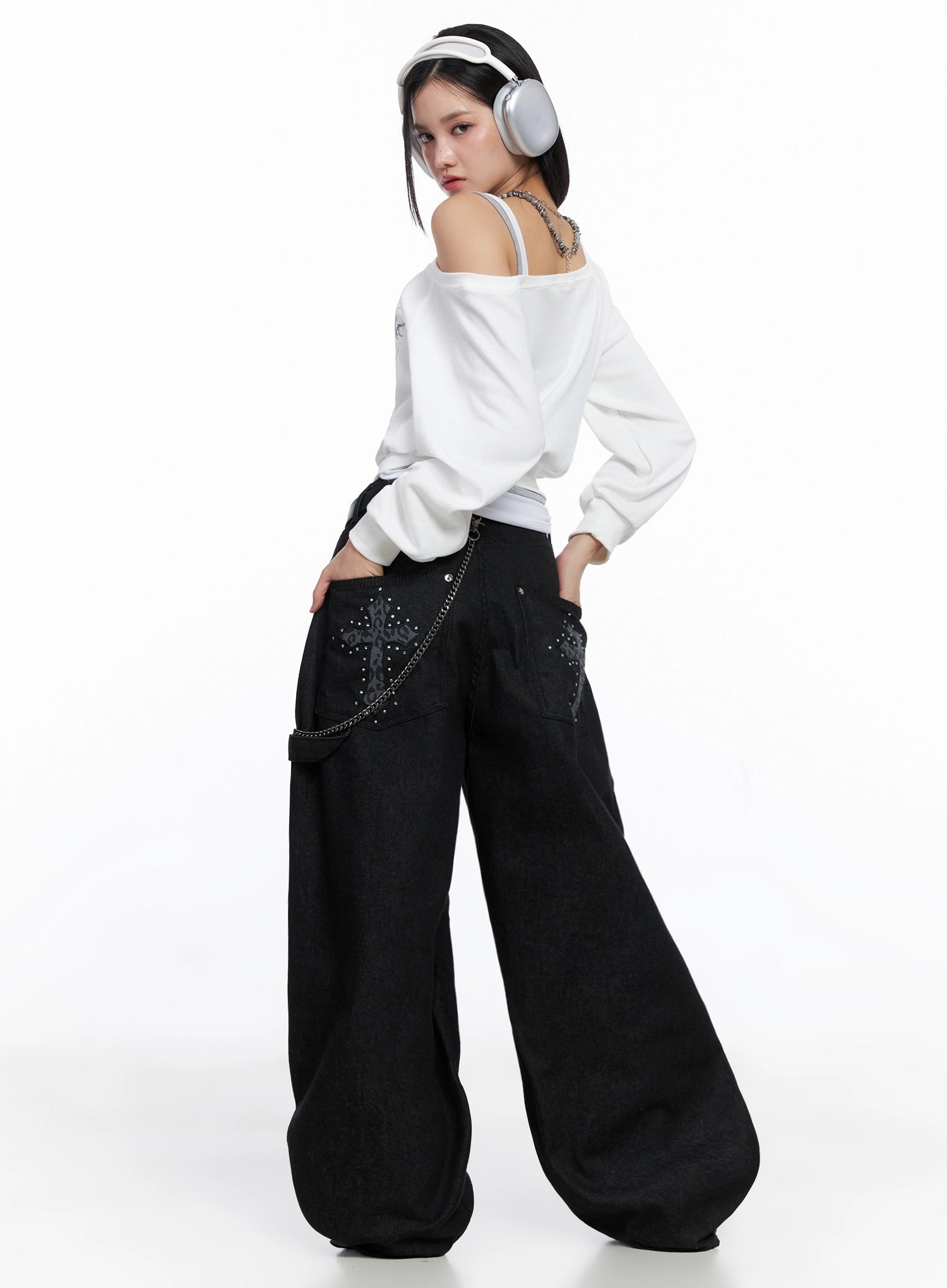 LEWKIN Lea Cross-Patch Baggy Jeans IS525 in black, back three-quarter view on model, ultra-wide leg cotton denim with stud-embellished leopard cross patch on pocket, chain detail, styled with white off-shoulder top.
