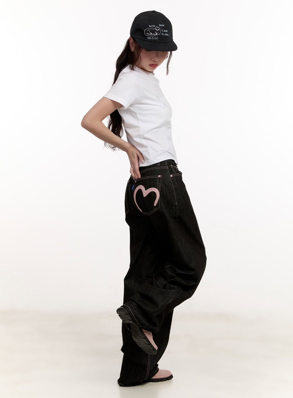 Trinity Stitched Baggy Jeans CM510 in black, side view on model, pink heart embroidery on back pocket visible, paired with white tee and black cap.