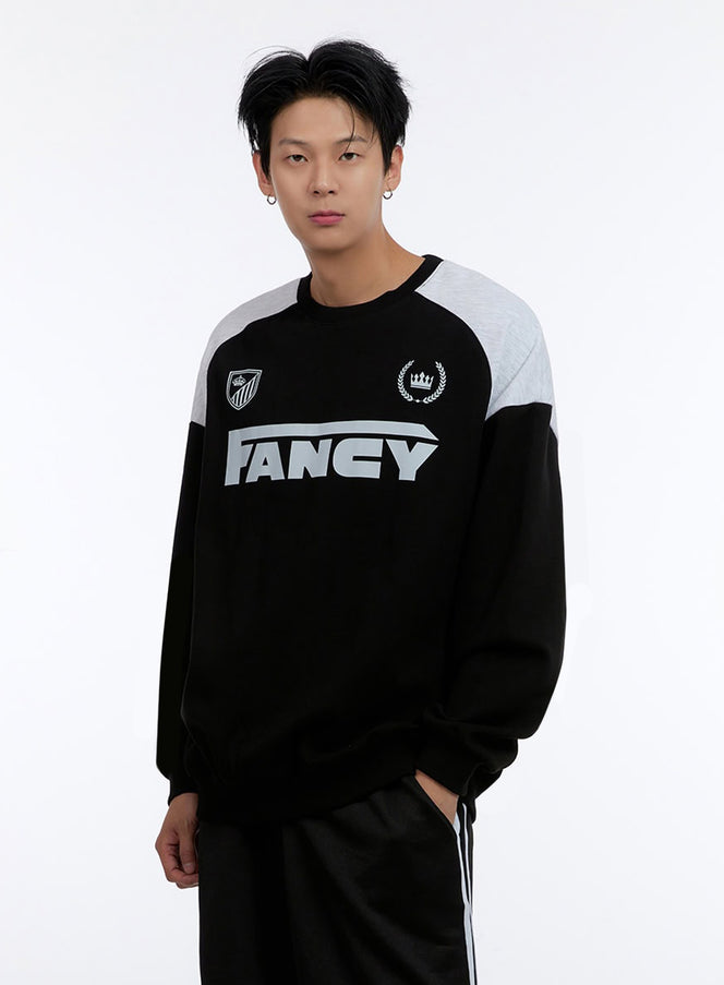 LEWKIN Men's Crew Neck Graphic Sweatshirt IO428 in Black, front three-quarter close-up on model, color-block grey raglan shoulders, FANCY logo print, shield and crown emblems, paired with black track pants.