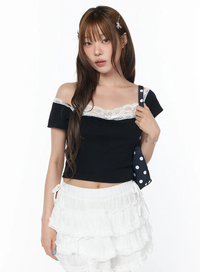 LEWKIN Off-Shoulder Lace Detail Crop Tee CY529 in black, front three-quarter shot on model holding polka-dot bag, white lace trim neckline, tight ribbed crop fit, styled with white tiered mini skirt.
