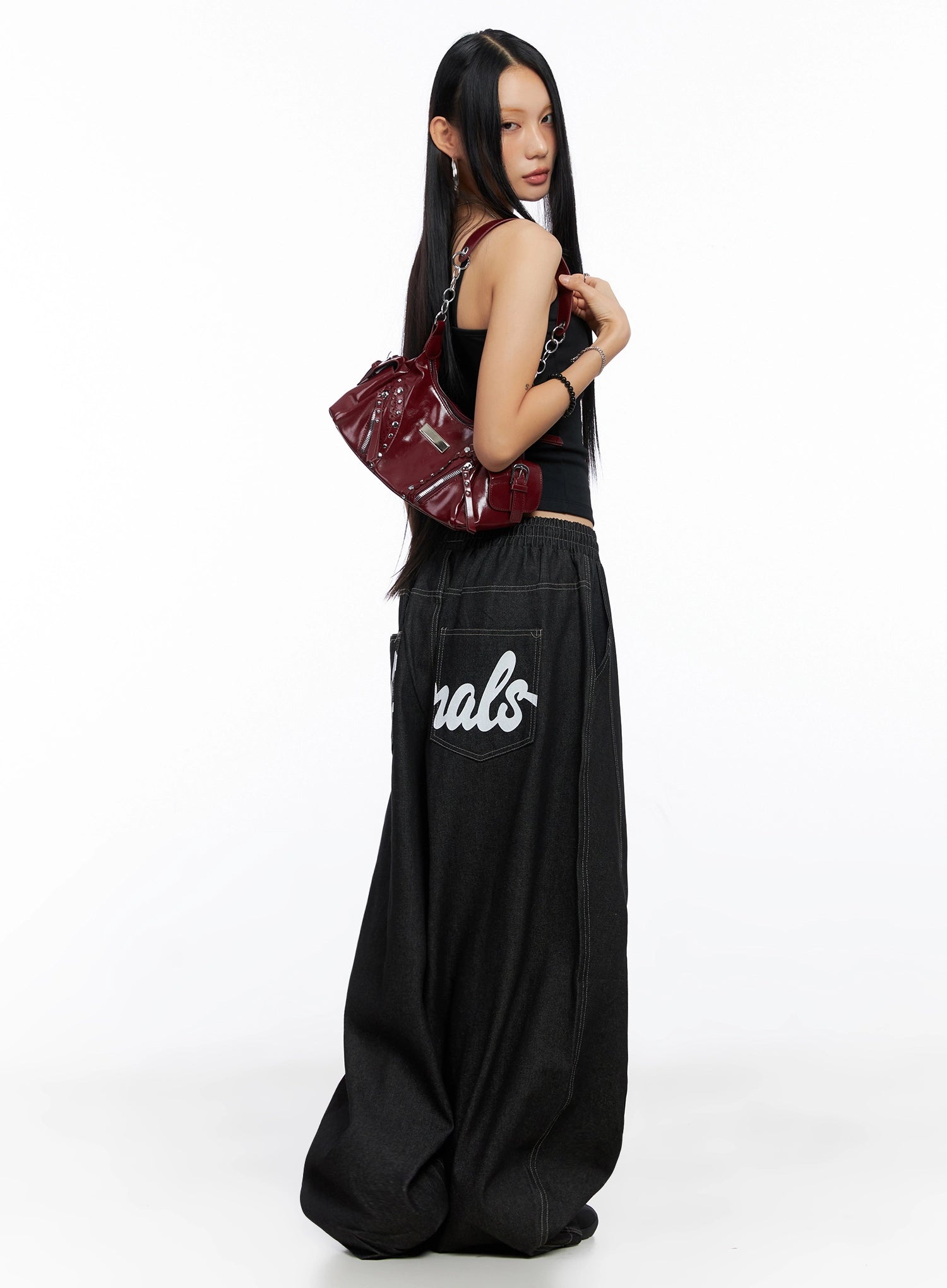 LEWKIN Ayra Graphic Baggy Jeans IO516 in Black, side view on model holding burgundy bag, ultra-wide cotton denim with cursive lettering on back pocket, paired with black cami top.