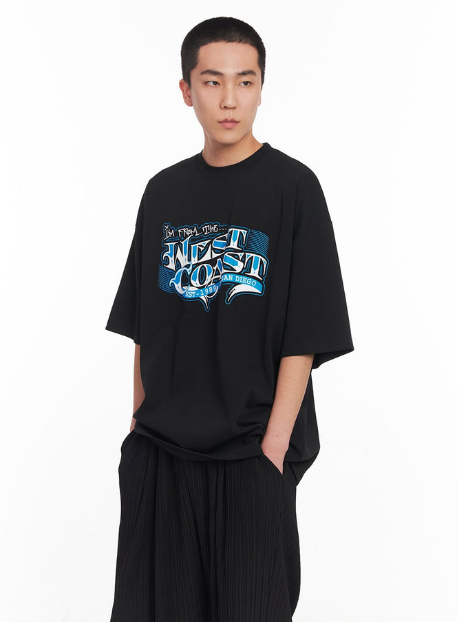 LEWKIN Men's West Coast Graphic Tee IY506 in black, angled front close-up on model, oversize cotton tee with blue graffiti-style chest graphic, paired with black pleated wide-leg trousers.
