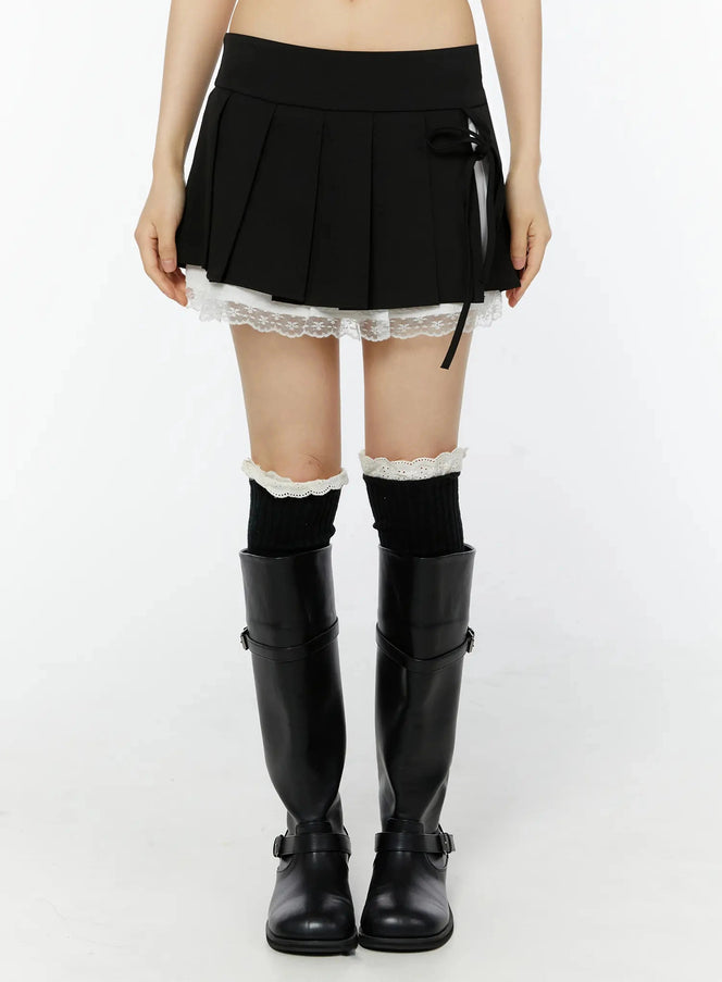 LEWKIN Chic Pleated Ribbon Lace Trim Mini Skirt IU518 in black, front waist-down shot on model, box pleats with white lace trim hem, side ribbon tie, paired with knee-high black boots.