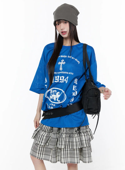LEWKIN Oversized Mesh Graphic Tee CU519 in Blue M/L, front three-quarter shot on model wearing grey beanie, white gothic cross and i1994 lion prints, mesh fabric, paired with plaid ruffle skirt and black shoulder bag.