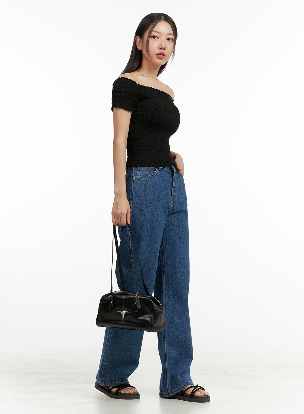 LEWKIN Blue Wide-Leg Jeans OU407 in blue, 3/4 side view on model holding black bag, wide-fit washed cotton denim with relaxed straight leg, styled with black off-shoulder top and sandals.