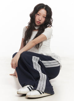 LEWKIN Angel Lace Wide-Leg Trackpants CA521 in dark blue, seated full-body editorial shot, model crouching with lace side stripe detail visible, paired with white lace top and sneakers.