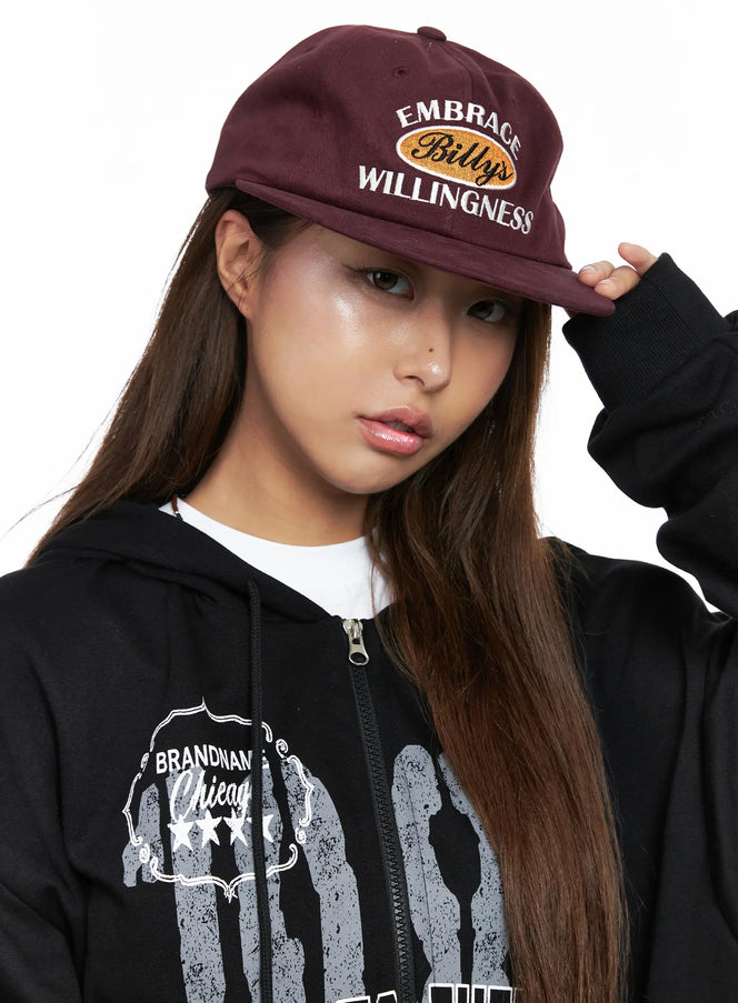 LEWKIN Embroidered Flat Cap CO513 in dark red, close-up front on model tilting brim, cotton with oval lettering embroidery, styled with black graphic zip hoodie.