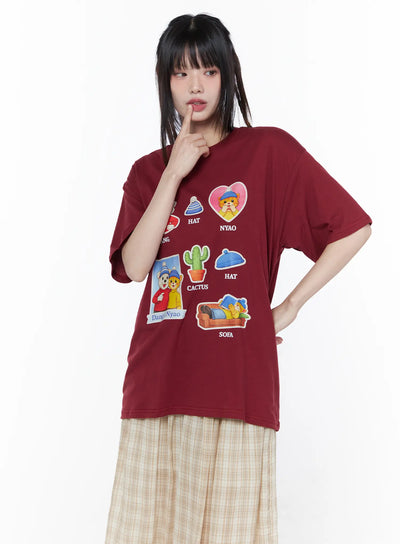 LEWKIN Oversized Graphic Print Tee CU519 in dark red, front three-quarter view on model, oversized cotton short sleeve with colorful Dang & Nyao cartoon sticker prints, paired with beige plaid midi skirt.