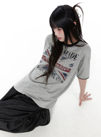 LEWKIN Graphic Short Sleeve Tee CU519 in gray, angled overhead shot on model seated on floor, distressed Union Jack print with black ringer trim, paired with dark wide-leg jeans.
