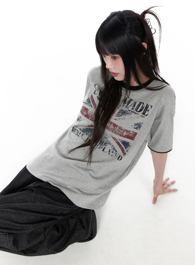 LEWKIN Graphic Short Sleeve Tee CU519 in gray, angled overhead shot on model seated on floor, distressed Union Jack print with black ringer trim, paired with dark wide-leg jeans.