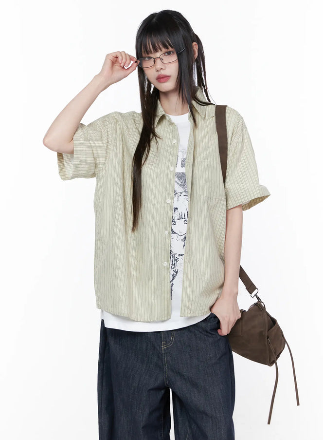 LEWKIN Oversized Short Sleeve Button-Up Shirt CU519 in light beige, front three-quarter shot on model adjusting glasses, striped woven fabric, worn open over graphic tee, paired with dark wide-leg jeans and brown bag.