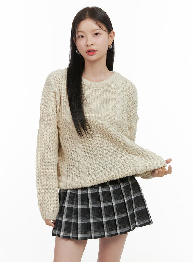 LEWKIN Basic Crew Neck Long Sleeve Knit OS411 in light beige, front three-quarter shot on model, oversize cable-knit sweater with round neck, paired with gray plaid pleated mini skirt.