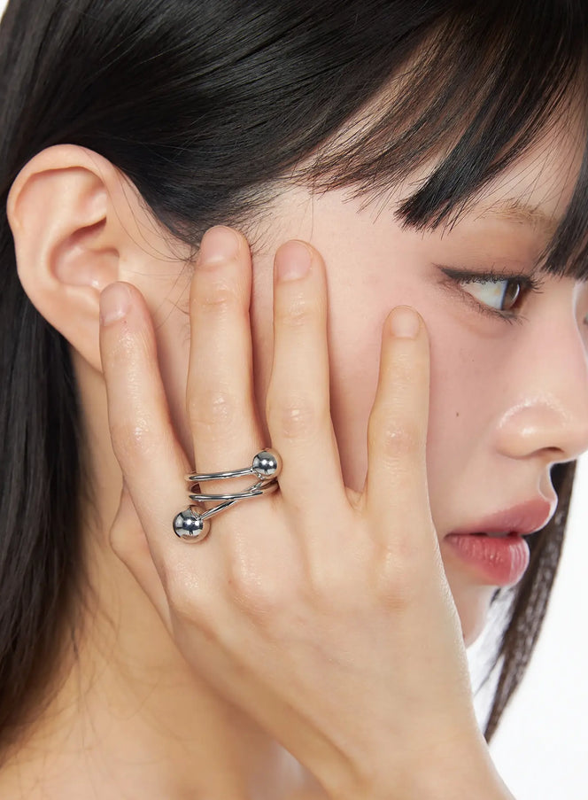 LEWKIN Orbital Twist Ring IY520 in silver, close-up on model's hand raised to face, triple-wire twist band with dual polished ball ends, glamorous K-pop style.