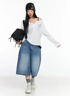 LEWKIN Oversized Cross Detail Long-Sleeve Top CA513 in White, full-body on model looking down, loose fit with lettering cross graphic, layered over black strap top, styled with wide-leg denim shorts, striped socks, and sneakers.