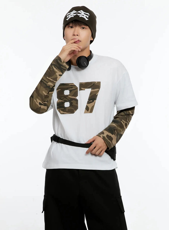 LEWKIN Men's Camo Graphic Layered Tee IN525 in white, front half-body on model with beanie and headphones, camo appliqué 87 graphic, layered camo long sleeves, styled with black cargo pants and belt bag.