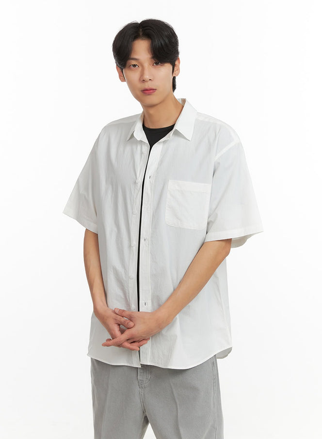 LEWKIN Men's Solid Buttoned Shirt (White) IY431 in white, front three-quarter view on model, oversized short-sleeve cotton shirt with chest patch pocket, layered over black tee, paired with grey trousers.