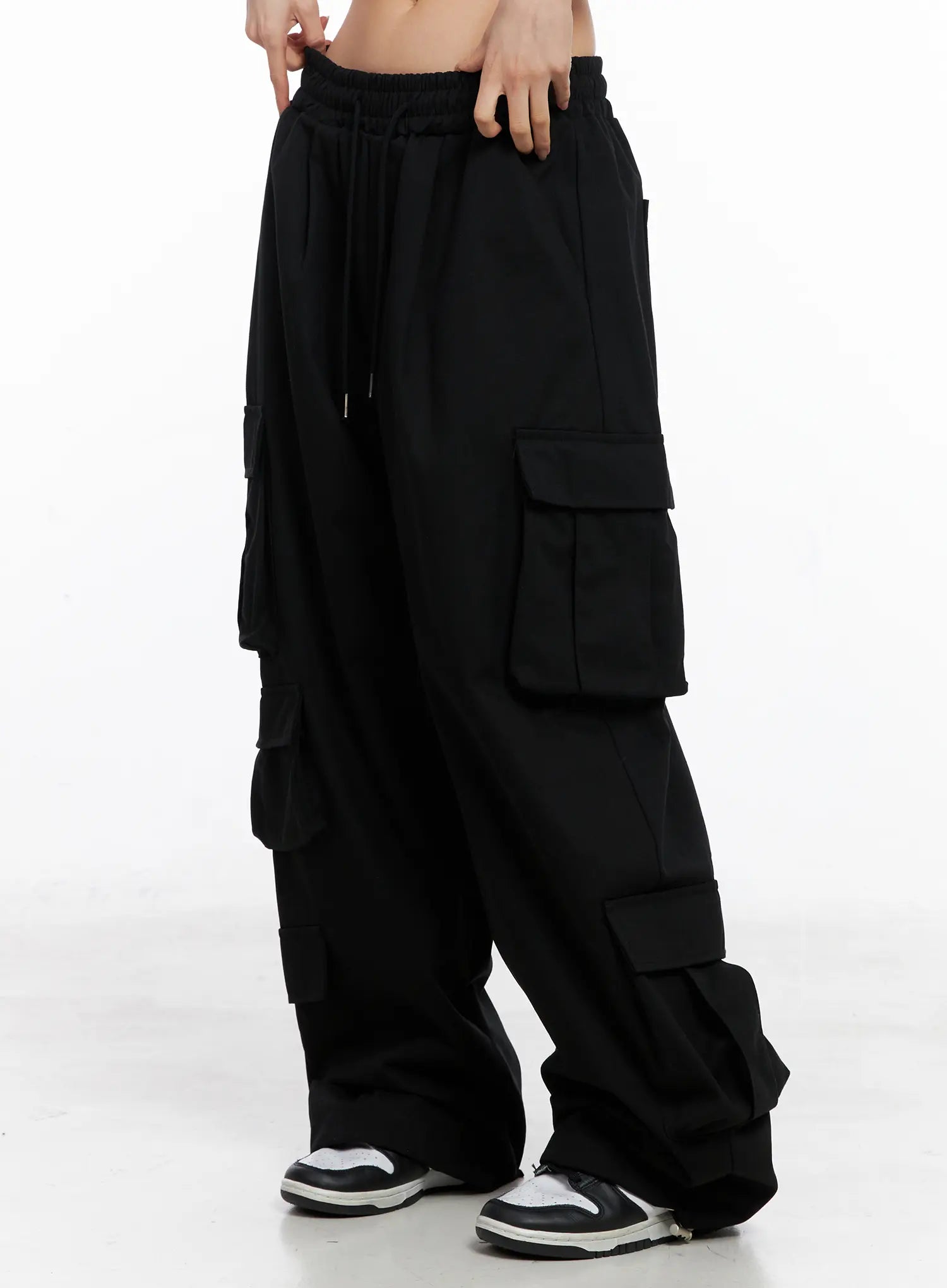 LEWKIN Wide Leg Cargo Pants CC1109 in black, side view on model, cotton wide-fit with elastic drawstring waist, multi-flap cargo pockets, long length, paired with white sneakers.