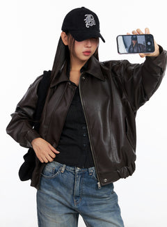 LEWKIN Faux Leather Bomber Jacket CD321 in Brown, front three-quarter shot on model taking selfie, zip-up collar, oversized polyester faux leather, styled with black top, cap, and blue jeans.