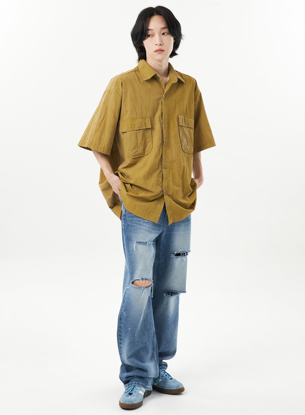 LEWKIN Ripped Jeans Unisex CU315 in mid blue, full-body front on male model, wide-leg cotton denim with heavy destroyed rips at thigh and knee, paint splatter, styled with olive short-sleeve shirt.