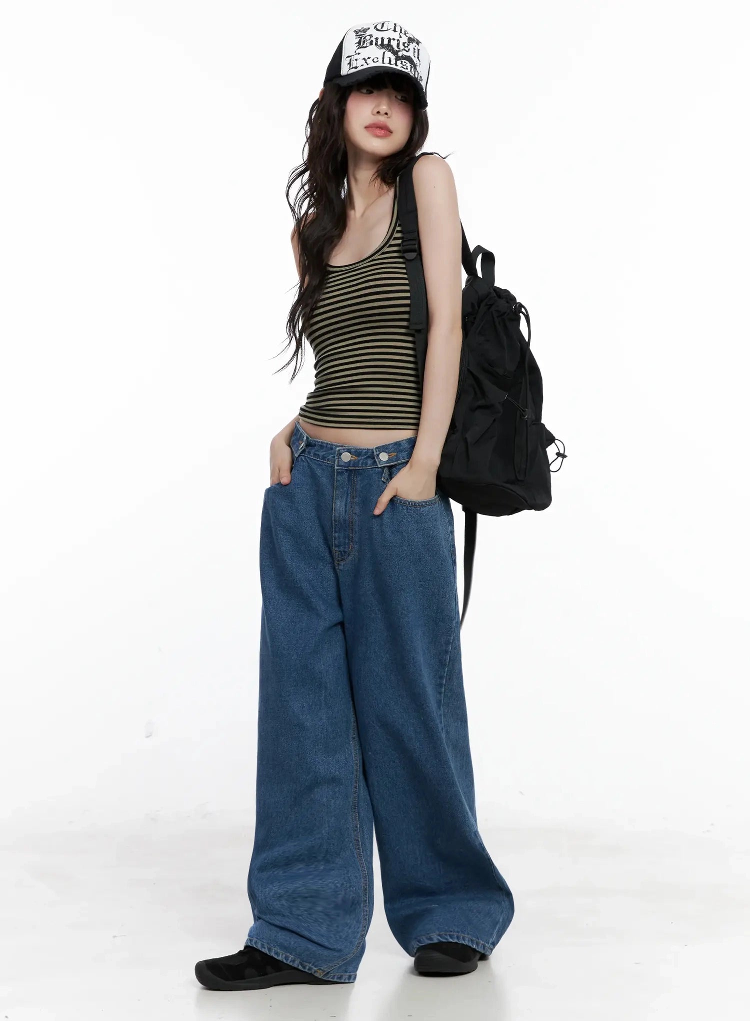 Pharita Loose-Fit Jeans IL528 in mid blue, full-body shot on model with trucker cap and backpack, long wide-leg silhouette paired with olive striped tank top, casual street style.