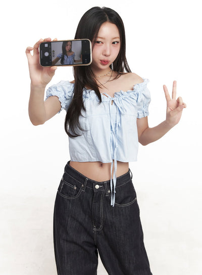 LEWKIN Ribbon Off-Shoulder Blouse Crop Top CA529 in light blue, fun selfie pose on model, puff sleeves with shirring, ribbon tie front detail, paired with dark wide-leg jeans.