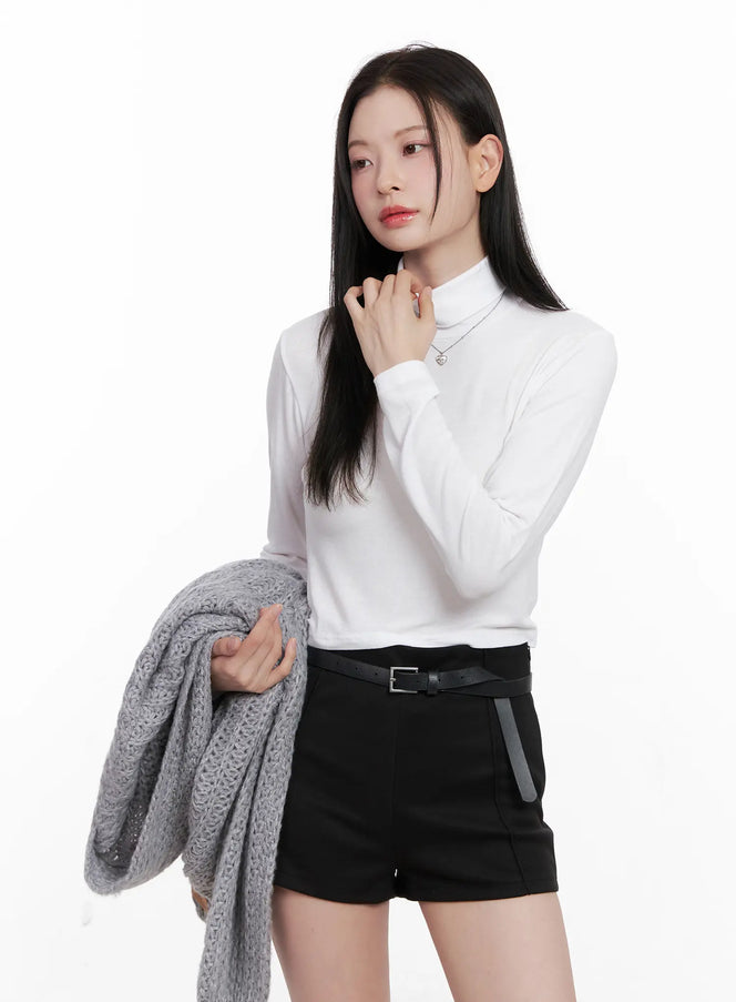 LEWKIN Crop Turtleneck Long Sleeve Top CN504 in white, front three-quarter shot on model holding grey knit cardigan, slim rayon-spandex crop silhouette, paired with black belted shorts.