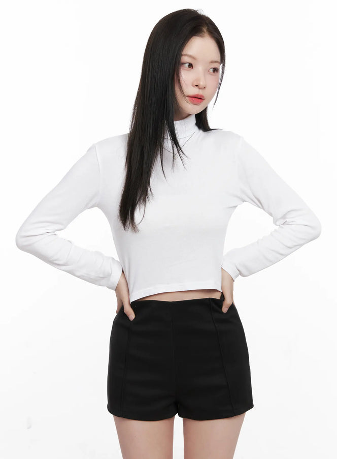 Crop Turtleneck Long Sleeve Top CN504 in white, front full-length shot on model hands on hips, cropped hem and fitted turtleneck visible, styled with black mini shorts.