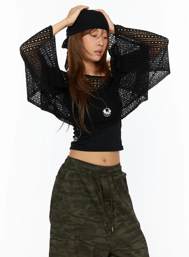 Boatneck Knit Long Sleeve Top CU517 in black, front waist-up shot, model arms raised, open-hole mesh knit sheer crop with wide sleeves over black tank, paired with camo cargo pants.