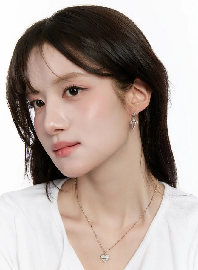 Elegant Cross Drop Earrings CM505 in silver, 3/4 face shot on model, crystal cross drop earring styled with white tee and delicate heart necklace, casual chic look.
