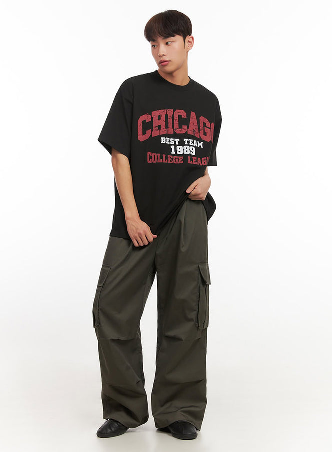 Men's Graphic T-Shirt IF521 in Black, full-body front shot on model, relaxed oversize silhouette with red varsity graphic, styled with wide-leg olive cargo trousers.
