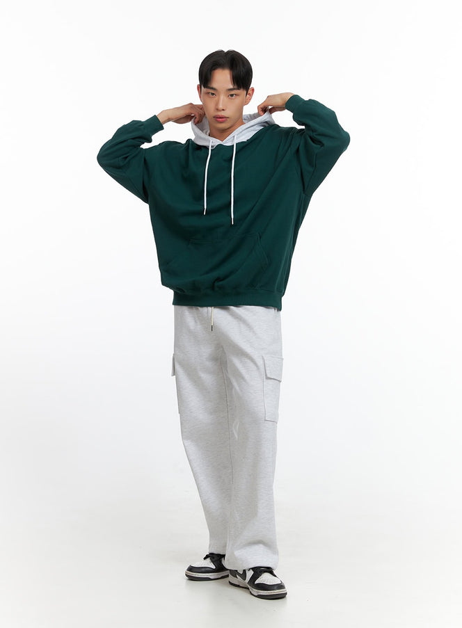 Men's Contrasting Hoodie (Green) IU414 in green, full-length front shot on model adjusting grey contrast hood, oversized fit, white drawstring, styled with grey cargo joggers and sneakers.