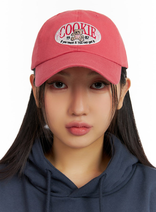 Bear Embroidered Graphic Cap IF421 in pink, front close-up on model, cotton baseball cap with oval bear embroidery and COOKIE lettering, paired with navy hoodie.