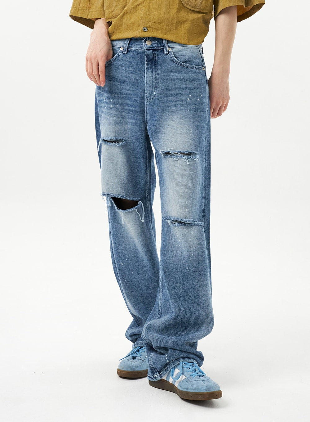 Ripped Jeans Unisex CU315 in mid blue, front close-up on model, wide-fit denim with frayed multi-rip detailing across thigh and knee, faded wash and paint splatter, paired with blue sneakers.