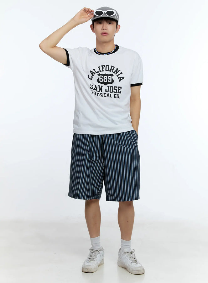 Men's Striped Straight-Leg Shorts IG515 in black, full-body front shot on male model wearing cap and graphic tee, relaxed straight-leg fit, multi-stripe woven fabric, styled with white sneakers.