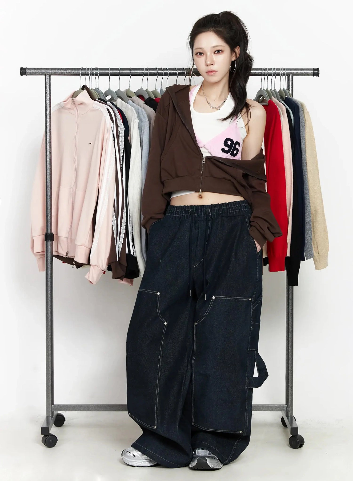 Ashlyn Carpenter Wide-Leg Jeans IJ528 in dark blue, full editorial shot, model posed by clothing rack, styled with brown zip hoodie and graphic tank, wide-leg silhouette with contrast stitching.
