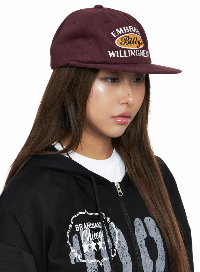 Embroidered Flat Cap CO513 in dark red, three-quarter front view on model, flat brim and structured crown, casual lettering embroidery, worn with black hoodie.