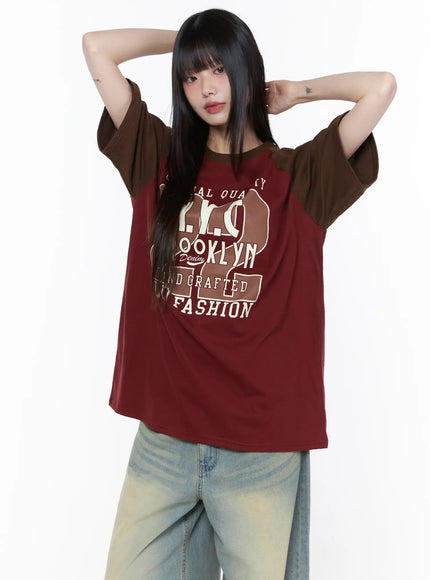 Casual Loose Fit Graphic Tee CU519 in wine red, front view on model arms raised, contrast brown raglan sleeves, oversized Brooklyn NYC graphic print, styled with baggy light-wash jeans.