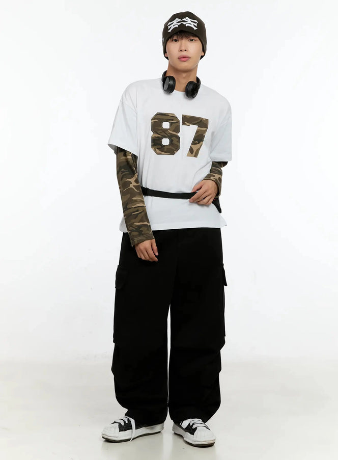 Men's Camo Graphic Layered Tee IN525 in white, full-body front on model, oversized fit with camo-print layered sleeves extending below short-sleeve hem, paired with black cargo pants and sneakers.