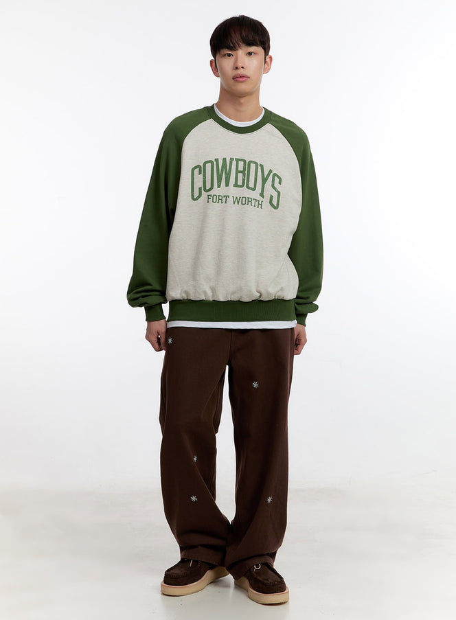 Men's Graphic Long Sleeve Raglan Sweatshirt IA504 in Green, full-body front shot on model, oversized fit with green raglan sleeves, Cowboys Fort Worth chest graphic, elastic hem, paired with brown wide-leg pants and loafers.