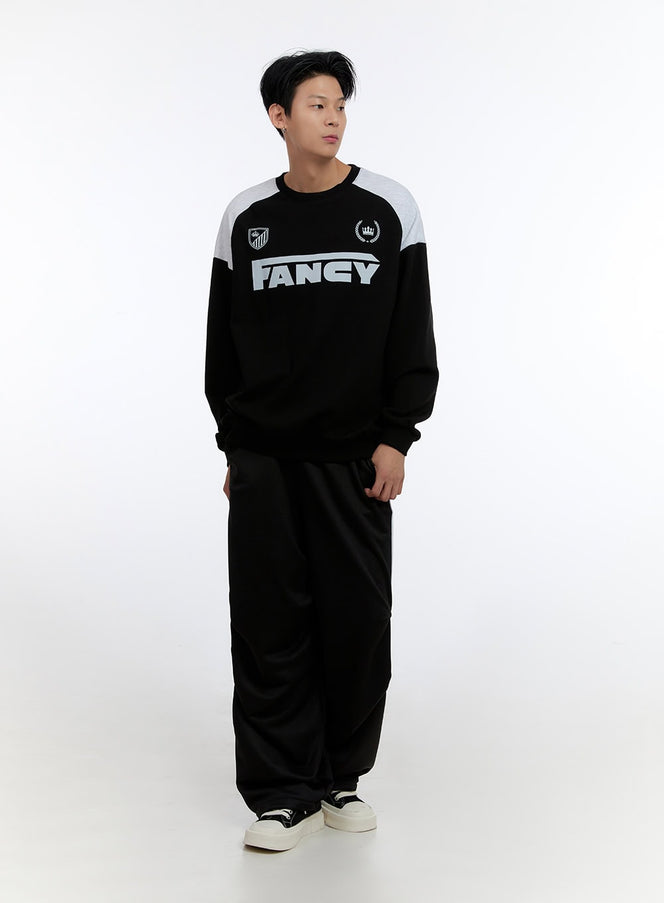 Men's Crew Neck Graphic Sweatshirt IO428 in Black, full-length front view on model, oversized fit with grey raglan color-block shoulders, FANCY graphic, styled with wide-leg black track pants and sneakers.