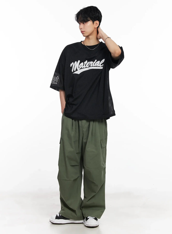 Men's Embroidered Mesh Graphic Tee IU526 in black, full-body front view on model with hand raised, mesh drop-shoulder silhouette, styled with green cargo trousers and sneakers.