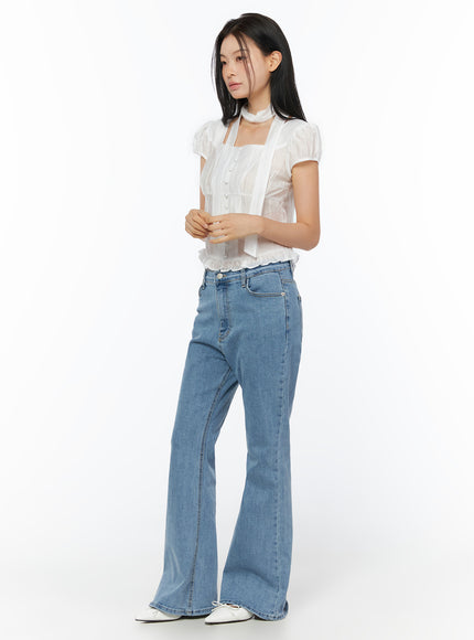 Leana Stretch Bootcut Jeans IU519 in blue, three-quarter side view on model, long bootcut silhouette, paired with white ruffle blouse and white flats.