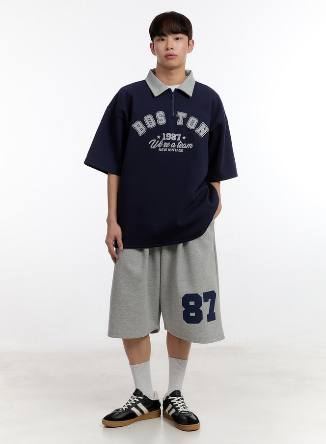 Men's Oversized Half-Zip Collared Sweatshirt IA504 in dark blue, full-body front on model, drop-shoulder short sleeves, varsity graphic, styled with grey sweat shorts and sneakers.