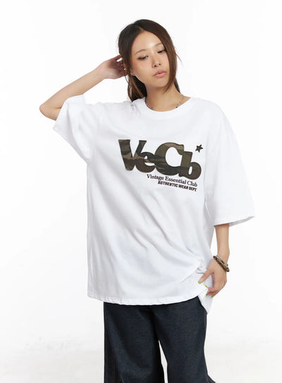 Oversized Camo Graphic Tee CM525 in white, front mid-shot on model, loose cotton round-neck tee featuring camo fabric patch VeCb logo, paired with dark wide-leg jeans.