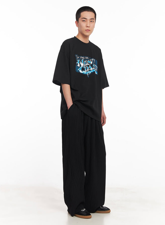 Men's West Coast Graphic Tee IY506 in black, three-quarter front angle full-length on model, drop-shoulder oversize fit showing hem length, styled with black wide-leg trousers and leather loafers.