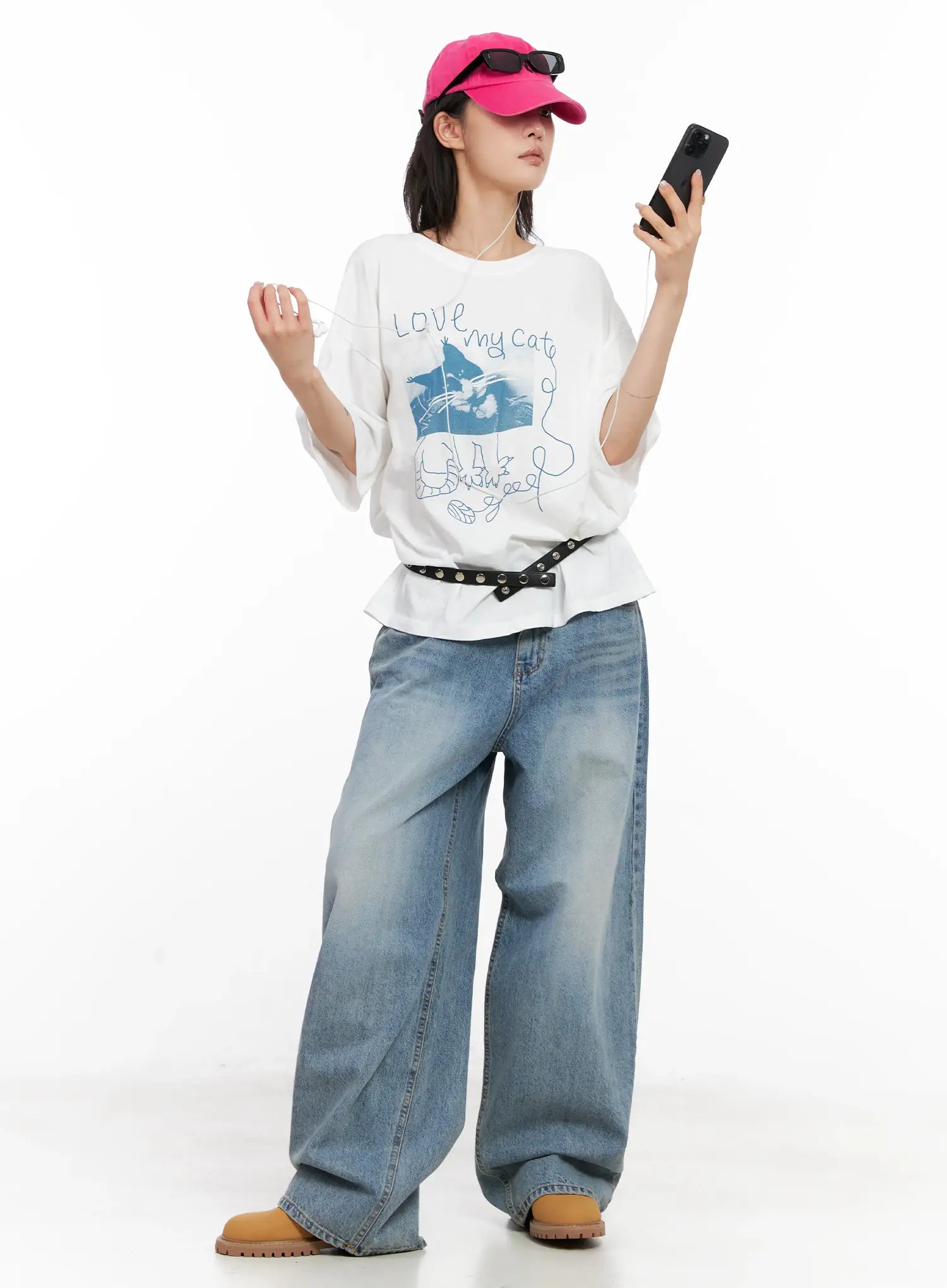 Koko Vintage-Wash Baggy Jeans CL528 in blue, full outfit shot on model holding phone, styled with white graphic tee, studded belt, and pink cap.