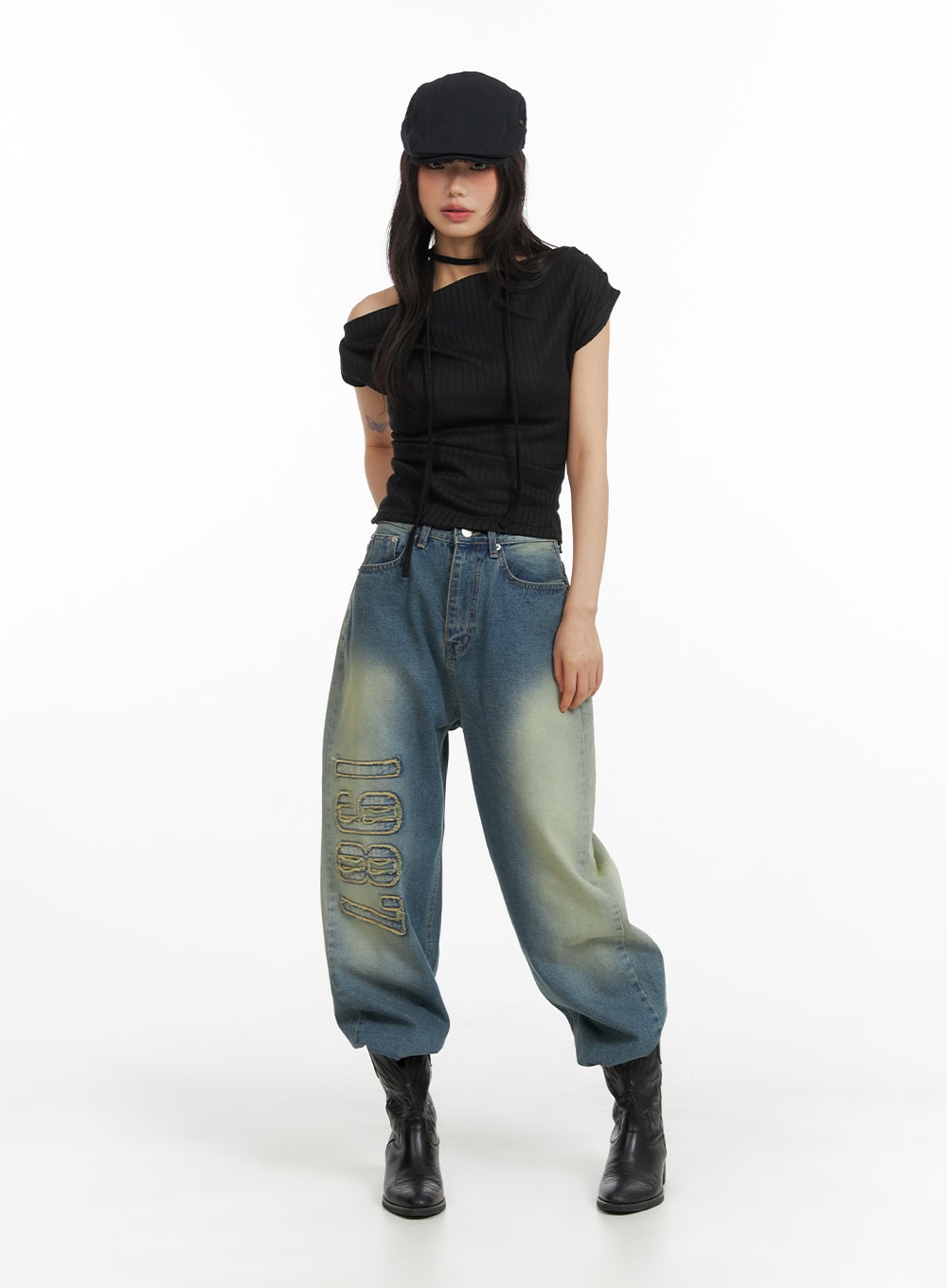 Distressed Baggy Straight Jeans CM413 in blue, full-body front on model, oversized baggy silhouette with yellow-tint wash and '1987' appliqué, styled with black asymmetric top, cap, and boots.