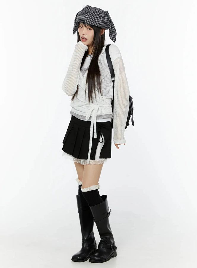 Chic Pleated Ribbon Lace Trim Mini Skirt IU518 in black, full-body side-angle shot on model, white lace underskirt peeking out, styled with cream knit wrap top, plaid bunny-ear hat, and knee-high boots.