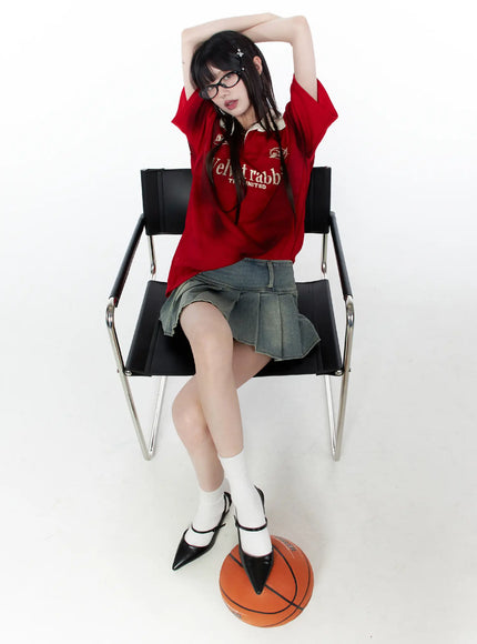 Oversized Collar Jersey CU519 in red, full-body seated on chair, oversized short sleeve with beige contrast collar and graphic print, styled with denim mini skirt, white socks, and heels, basketball at feet.